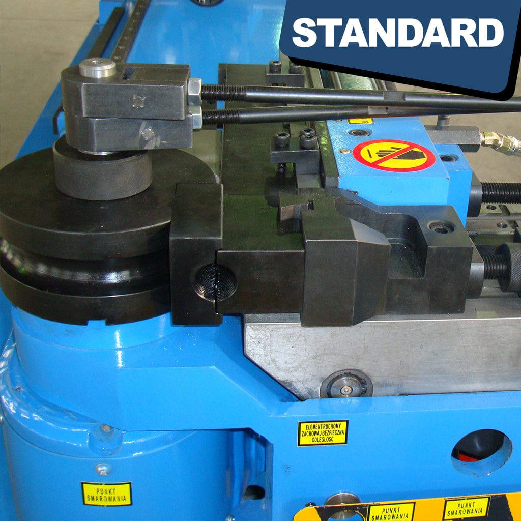 A hydraulic mandrel tube bender in action, bending a metal tube. The machine, labeled STANDARD BTNC-129, shows a hydraulic arm bending a cylindrical metal tube around a central axis. The tube passes through various rollers and bending mechanisms, displaying the industrial process of tube bending.