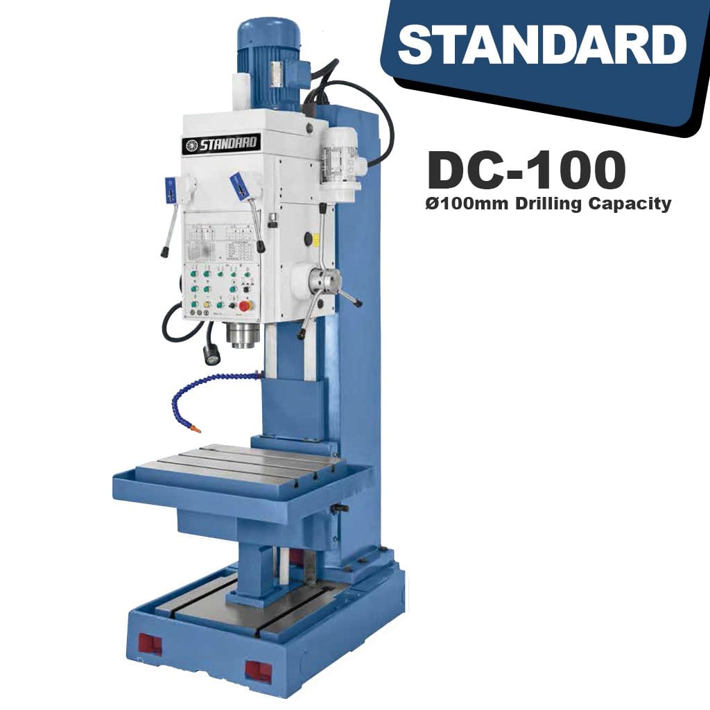 An image of the STANDARD DC-100 Heavy Duty Column Type Pedestal Drilling Machine. The machine is large and metallic, standing upright with a vertical column and a drilling apparatus at the top. Various knobs, handles, and controls are visible on its surface.