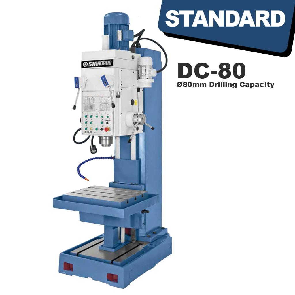  A sturdy, grey STANDARD DC-80 Heavy Duty Column Type Pedestal Drilling Machine. The machine stands tall with a robust column and a large work table. Various controls and a powerful drilling mechanism are visible, indicating its heavy-duty construction and industrial functionality.
